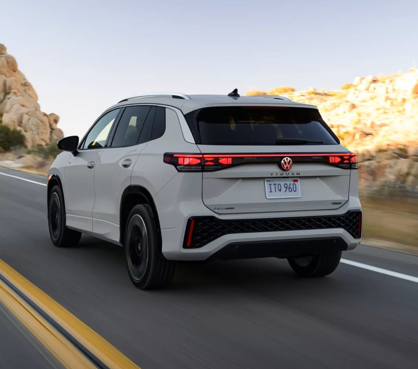 Rear three-quarter view of a white Volkswagen Tiguan driving on a paved road, highlighting its full-width LED taillight bar, sculpted rear design, and black alloy wheels.
