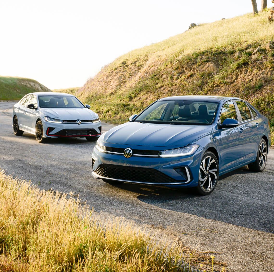 Front three-quarter view of blue Volkswagen Jetta SEL and white Jetta GLI on a rural road by a grassy hill.
