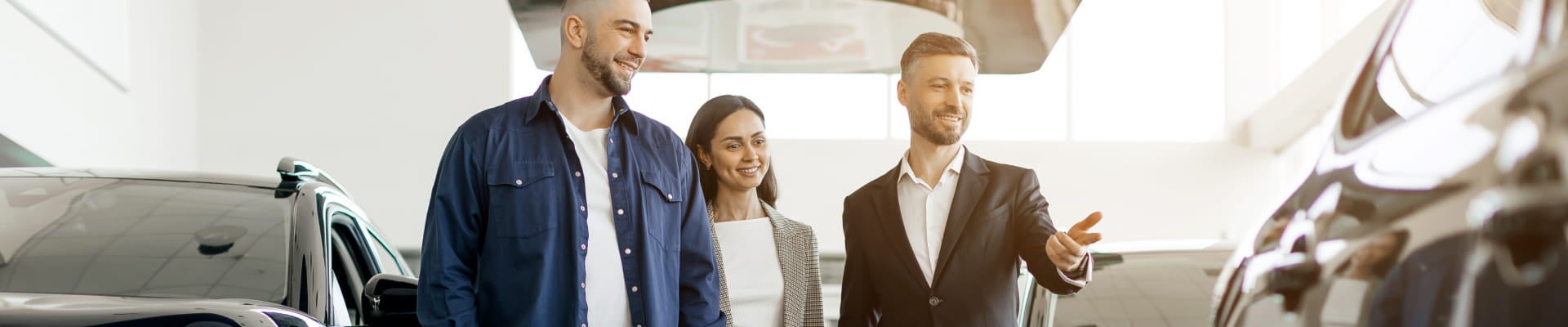 Couple speaking with a salesperson while looking at vehicles inside a dealership showroom.